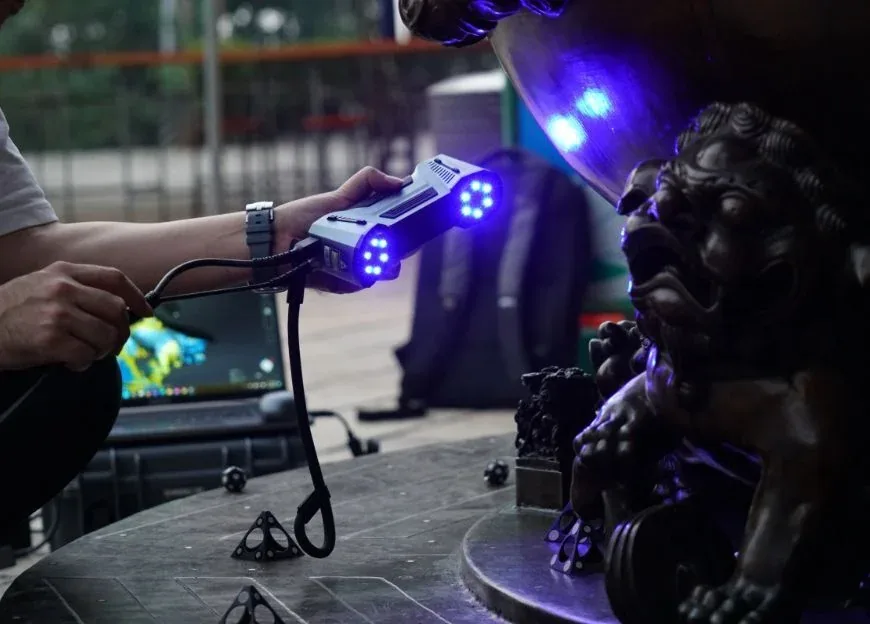 Un opérateur numérise une sculpture de lion en bronze avec un scanner 3D à main ; les lignes laser bleues balaient la surface, le modèle se construit sur l'écran d'un ordinateur portable.